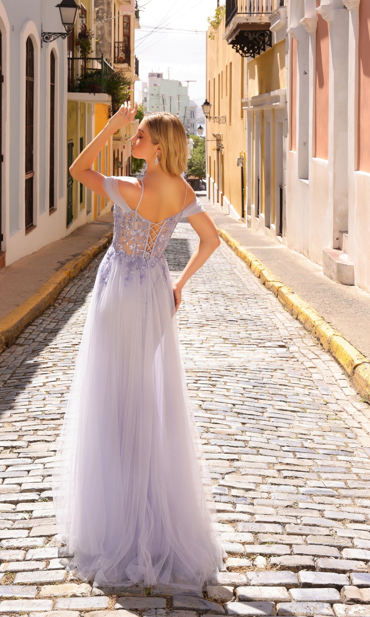 A woman in the Nox Anabel L1362 Overskirt Long Sequin Prom Dress stands on a sunny cobblestone street, facing away with her hand to her forehead.